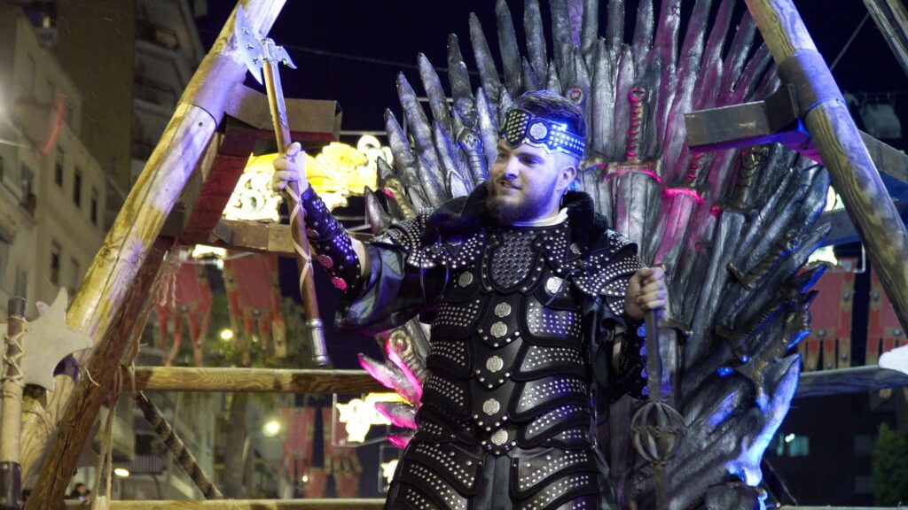 Rey con armadura oscura sentado en un trono durante el desfile nocturno.