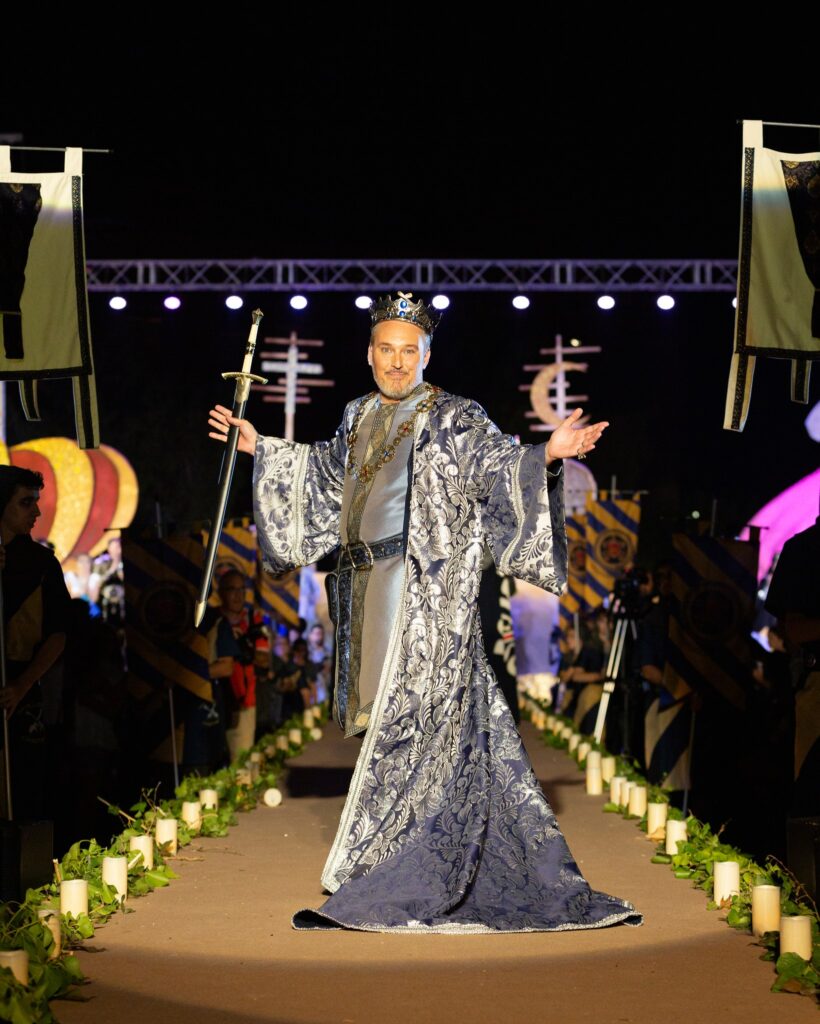 Rey con corona, espada y manto azul y plata sobre una pasarela iluminada con velas.